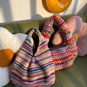 K&ouml;nnte beinhalten: Zwei handgefertigte H&auml;keltaschen in Regenbogenfarben. Eine Tasche hat eine runde Form mit einem einzelnen Riemen, die andere eine strukturiertere Form mit einem k&uuml;rzeren Riemen. Die Taschen sind aus farbenfrohem Garn gefertigt.