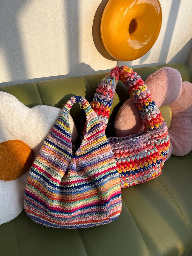 K&ouml;nnte beinhalten: Zwei handgefertigte H&auml;keltaschen in Regenbogenfarben. Eine Tasche hat eine runde Form mit einem einzelnen Riemen, die andere eine strukturiertere Form mit einem k&uuml;rzeren Riemen. Die Taschen sind aus farbenfrohem Garn gefertigt.