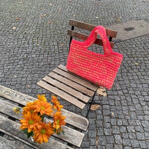 May include: A pink and yellow crocheted tote bag with a wooden handle sits on a wooden bench. The bench is outdoors on a stone patio.