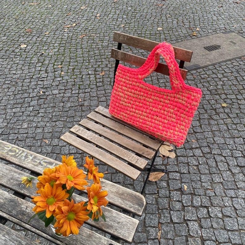 May include: A pink and yellow crocheted tote bag with a wooden handle sits on a wooden bench. The bench is outdoors on a stone patio.