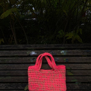 May include: A pink and beige crocheted tote bag with a single handle on a wooden bench.