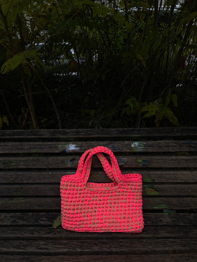May include: A pink and beige crocheted tote bag with a single handle on a wooden bench.