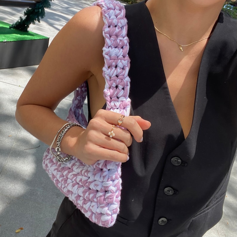 May include: A pink and white crocheted tote bag with a thick strap. The bag is being held by a person wearing a black waistcoat.