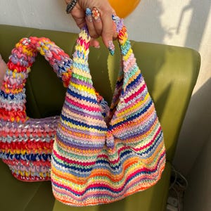 K&ouml;nnte beinhalten: Zwei farbenfrohe, handgefertigte H&auml;keltaschen. Eine ist eine kleine, dreieckige Handtasche mit einem Regenbogen aus horizontalen Streifen. Die andere ist eine gr&ouml;&szlig;ere, runde Tasche mit einem &auml;hnlichen Farbschema. Beide Taschen haben ein einzigartiges, strukturiertes Aussehen.