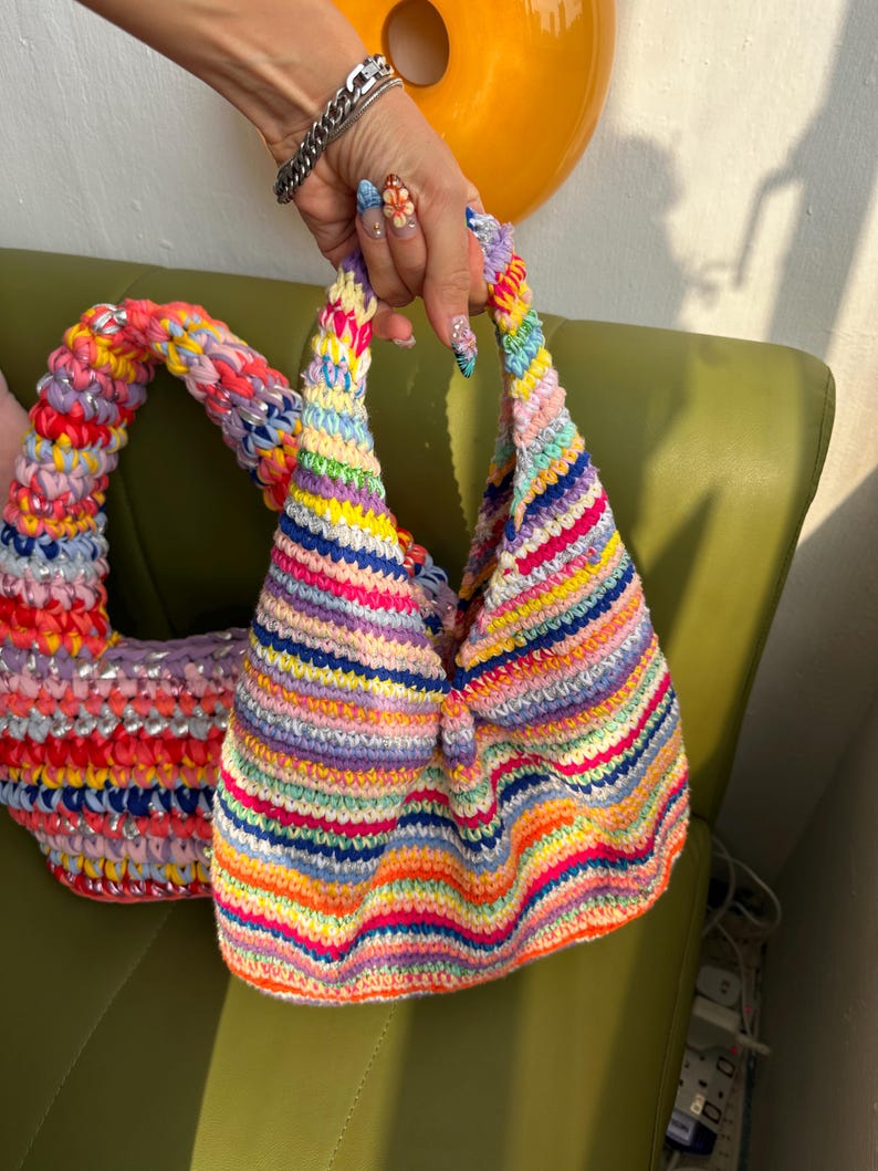 K&ouml;nnte beinhalten: Zwei farbenfrohe, handgefertigte H&auml;keltaschen. Eine ist eine kleine, dreieckige Handtasche mit einem Regenbogen aus horizontalen Streifen. Die andere ist eine gr&ouml;&szlig;ere, runde Tasche mit einem &auml;hnlichen Farbschema. Beide Taschen haben ein einzigartiges, strukturiertes Aussehen.