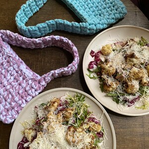 May include: Two crocheted bags, one light blue and one purple, are on a wooden table with two plates of salad. The salad is made with roasted cauliflower, rocket, and parmesan cheese.