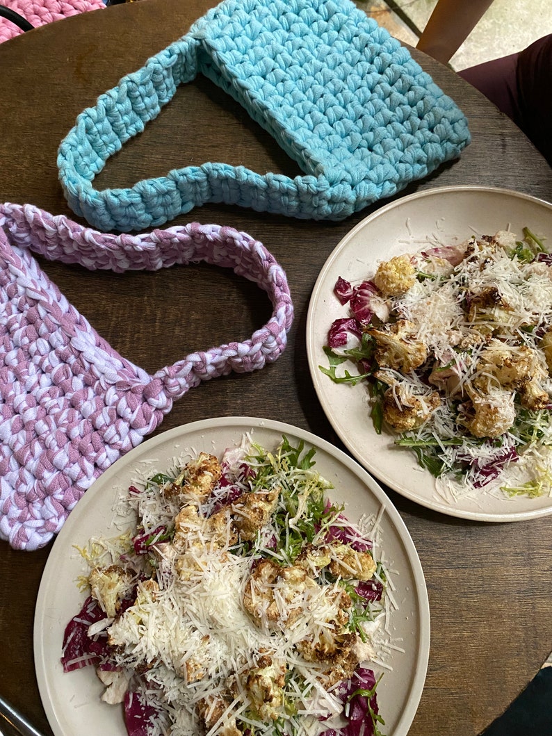 May include: Two crocheted bags, one light blue and one purple, are on a wooden table with two plates of salad. The salad is made with roasted cauliflower, rocket, and parmesan cheese.