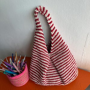 Puede incluir: Un bolso de ganchillo hecho a mano con rayas horizontales rojas y blancas. El bolso tiene forma triangular y un asa a juego a rayas. Una cesta rosa llena de ganchos de ganchillo est&aacute; junto al bolso sobre una mesa naranja.