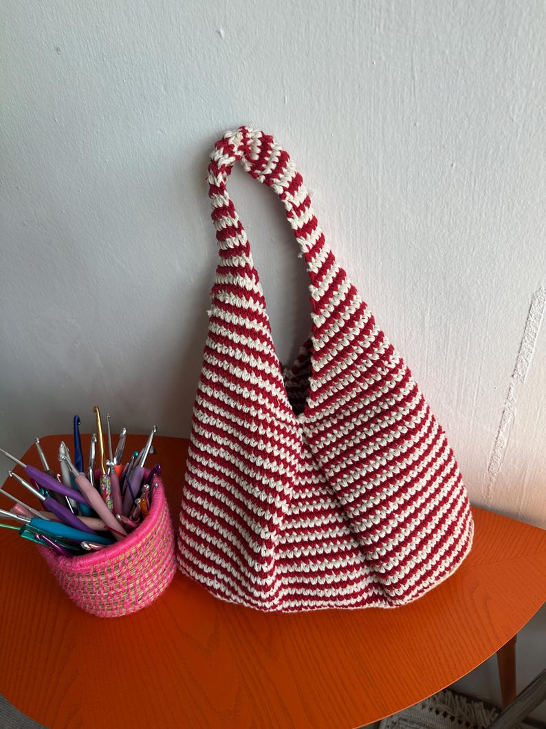Puede incluir: Un bolso de ganchillo hecho a mano con rayas horizontales rojas y blancas. El bolso tiene forma triangular y un asa a juego a rayas. Una cesta rosa llena de ganchos de ganchillo est&aacute; junto al bolso sobre una mesa naranja.
