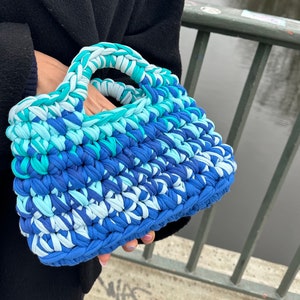 May include: A blue, white, and green crocheted tote bag with a single handle. The bag is made of thick yarn and has a textured surface.