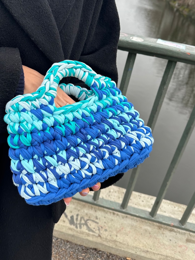 May include: A blue, white, and green crocheted tote bag with a single handle. The bag is made of thick yarn and has a textured surface.