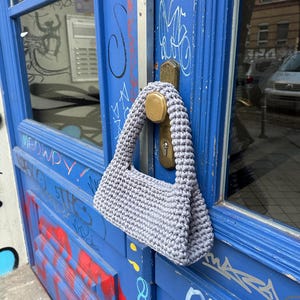 Pu&ograve; includere: Una borsa a tracolla grigia, lavorata all'uncinetto a mano, di forma rettangolare e con un manico spesso e intrecciato. La borsa &egrave; appesa a una porta blu con graffiti e una maniglia in ottone. La trama della borsa &egrave; visibile.