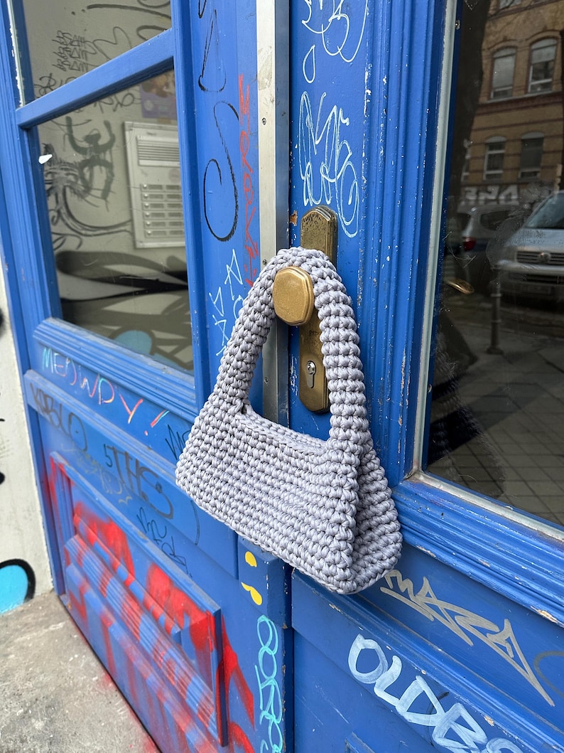 Pu&ograve; includere: Una borsa a tracolla grigia, lavorata all'uncinetto a mano, di forma rettangolare e con un manico spesso e intrecciato. La borsa &egrave; appesa a una porta blu con graffiti e una maniglia in ottone. La trama della borsa &egrave; visibile.