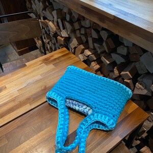 May include: A turquoise blue crocheted tote bag with light blue handles. The bag is sitting on a wooden table.