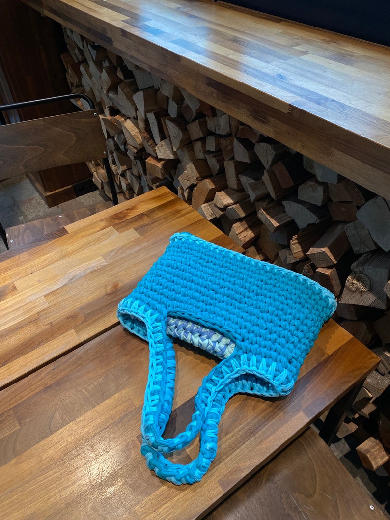 May include: A turquoise blue crocheted tote bag with light blue handles. The bag is sitting on a wooden table.