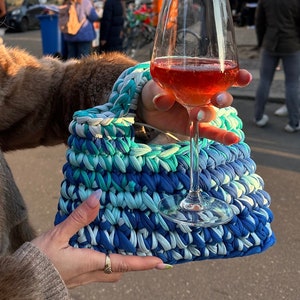 May include: A blue and green crocheted bag with a white glass of wine in front of it.