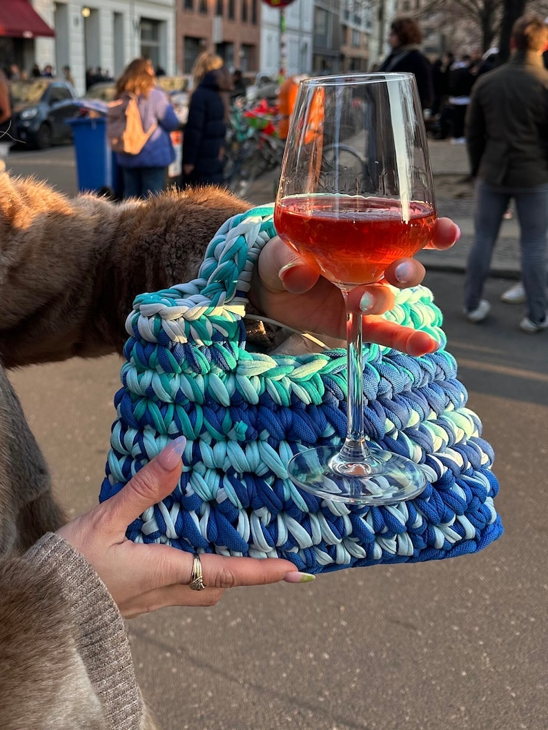 May include: A blue and green crocheted bag with a white glass of wine in front of it.