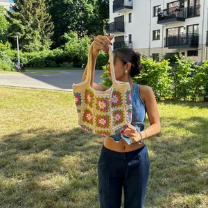 Op de afbeelding: Een vrouw houdt een gehaakte tote bag vast met een bloemenpatroon in roze, groene en gele tinten. De tas heeft een cr&egrave;mekleurige achtergrond en is gemaakt van vierkante granny stitch blokken.