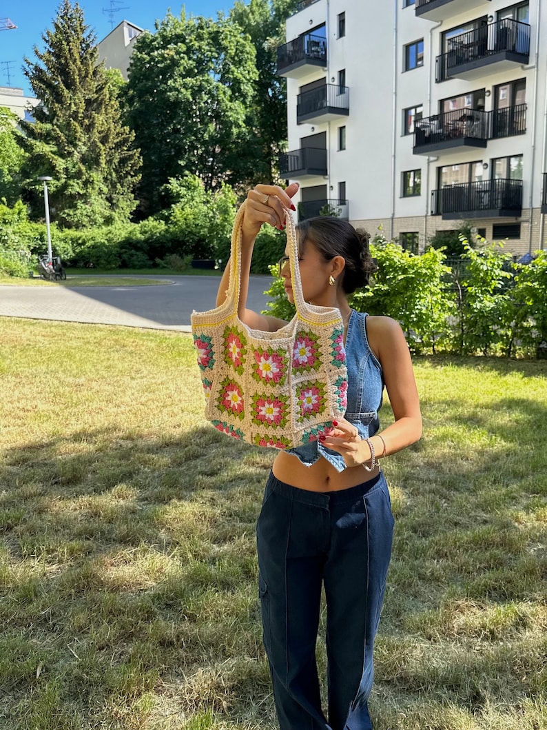Op de afbeelding: Een vrouw houdt een gehaakte tote bag vast met een bloemenpatroon in roze, groene en gele tinten. De tas heeft een cr&egrave;mekleurige achtergrond en is gemaakt van vierkante granny stitch blokken.