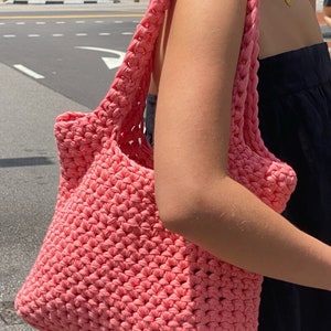 May include: A pink crocheted tote bag with a wide, open top and thick straps. The bag is made of a soft, textured yarn and has a casual, relaxed style.