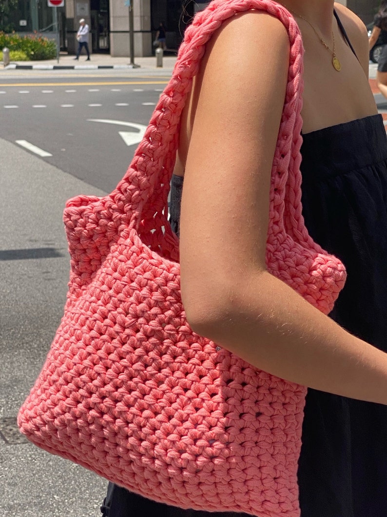 May include: A pink crocheted tote bag with a wide, open top and thick straps. The bag is made of a soft, textured yarn and has a casual, relaxed style.