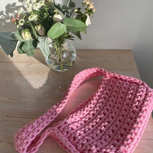 K&ouml;nnte beinhalten: Eine rosafarbene geh&auml;kelte Tragetasche mit einem langen Riemen. Die Tasche ist aus dickem Garn gefertigt und hat ein schlichtes, l&auml;ssiges Design.