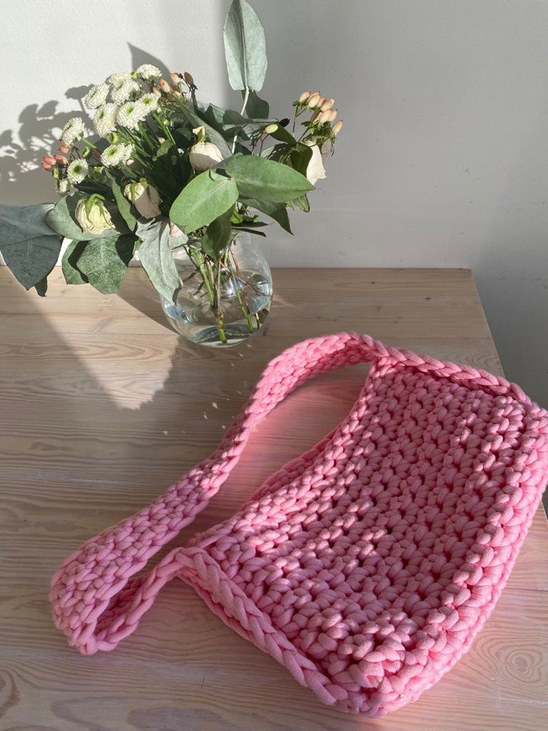 K&ouml;nnte beinhalten: Eine rosafarbene geh&auml;kelte Tragetasche mit einem langen Riemen. Die Tasche ist aus dickem Garn gefertigt und hat ein schlichtes, l&auml;ssiges Design.