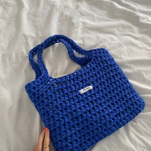 May include: A blue crocheted tote bag with a white label that says "BYOAK." The bag is made with a thick, textured yarn and has a simple, open weave.