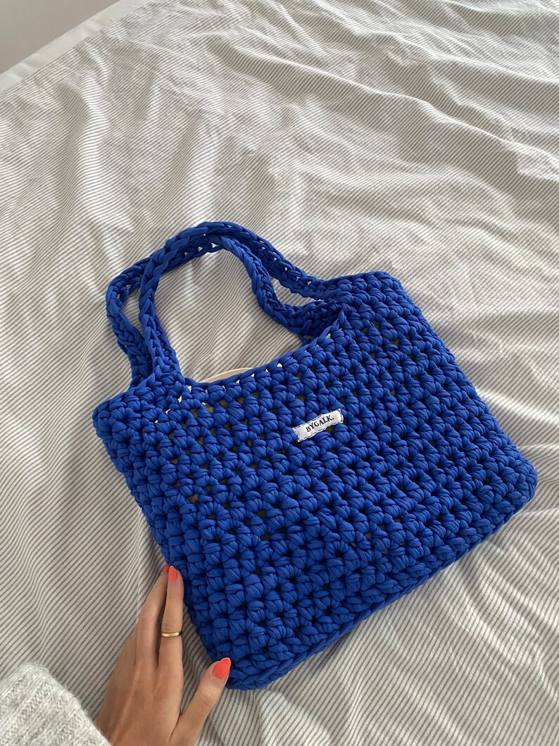 May include: A blue crocheted tote bag with a white label that says "BYOAK." The bag is made with a thick, textured yarn and has a simple, open weave.