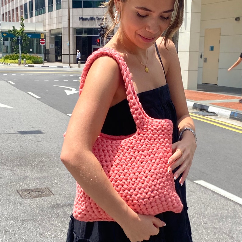 May include: A pink crocheted tote bag with a thick, textured weave. The bag has a single strap and is being held by a person.