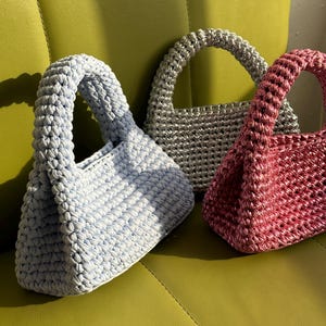Pu&ograve; includere: Tre borse all'uncinetto fatte a mano in colori pastello: azzurro, argento e rosa. Ogni borsa ha un manico arrotondato e un aspetto intrecciato strutturato. Le borse sono esposte su una superficie verde, mostrando la loro lavorazione unica.