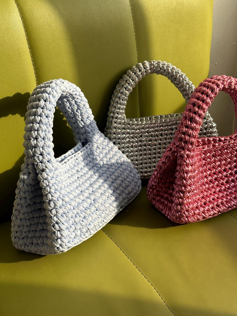 Pu&ograve; includere: Tre borse all'uncinetto fatte a mano in colori pastello: azzurro, argento e rosa. Ogni borsa ha un manico arrotondato e un aspetto intrecciato strutturato. Le borse sono esposte su una superficie verde, mostrando la loro lavorazione unica.
