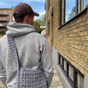 K&ouml;nnte beinhalten: Eine Person tr&auml;gt einen grauen Hoodie und eine braune Baseballkappe und tr&auml;gt eine graue geh&auml;kelte Umh&auml;ngetasche.