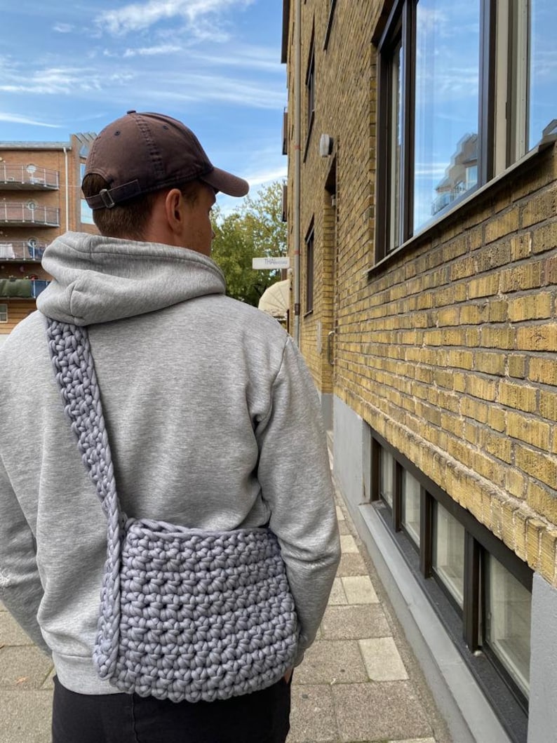K&ouml;nnte beinhalten: Eine Person tr&auml;gt einen grauen Hoodie und eine braune Baseballkappe und tr&auml;gt eine graue geh&auml;kelte Umh&auml;ngetasche.