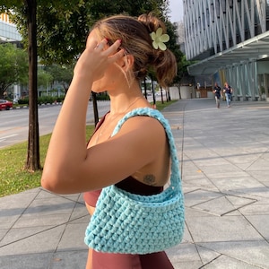 May include: A light blue crocheted handbag with a single strap. The bag is being worn by a person with a flower hair clip in their hair.