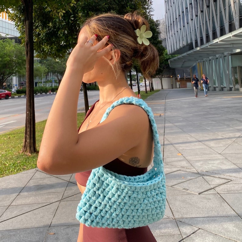 May include: A light blue crocheted handbag with a single strap. The bag is being worn by a person with a flower hair clip in their hair.