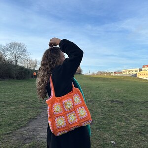 Op de afbeelding: Een vrouw draagt een feloranje gehaakte tote bag met roze en gele bloemen. De tas is gemaakt van granny squares.