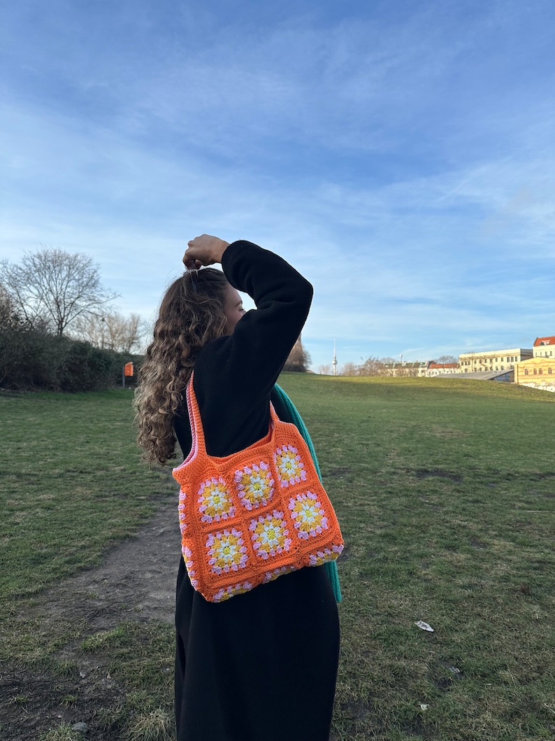Op de afbeelding: Een vrouw draagt een feloranje gehaakte tote bag met roze en gele bloemen. De tas is gemaakt van granny squares.