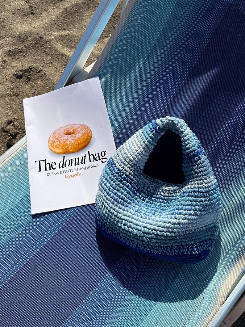 Może przedstawiać: Niebiesko-biała szydełkowa torebka leży na niebieskim krześle plażowym w paski obok białej papierowej torby. Na torbie widnieje napis "The donut bag" i zdjęcie pączka.
