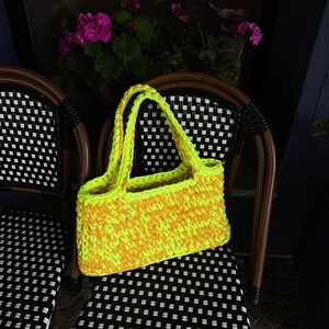 May include: A bright yellow and orange crocheted tote bag with thick straps. The bag is sitting on a black and white checkered chair.