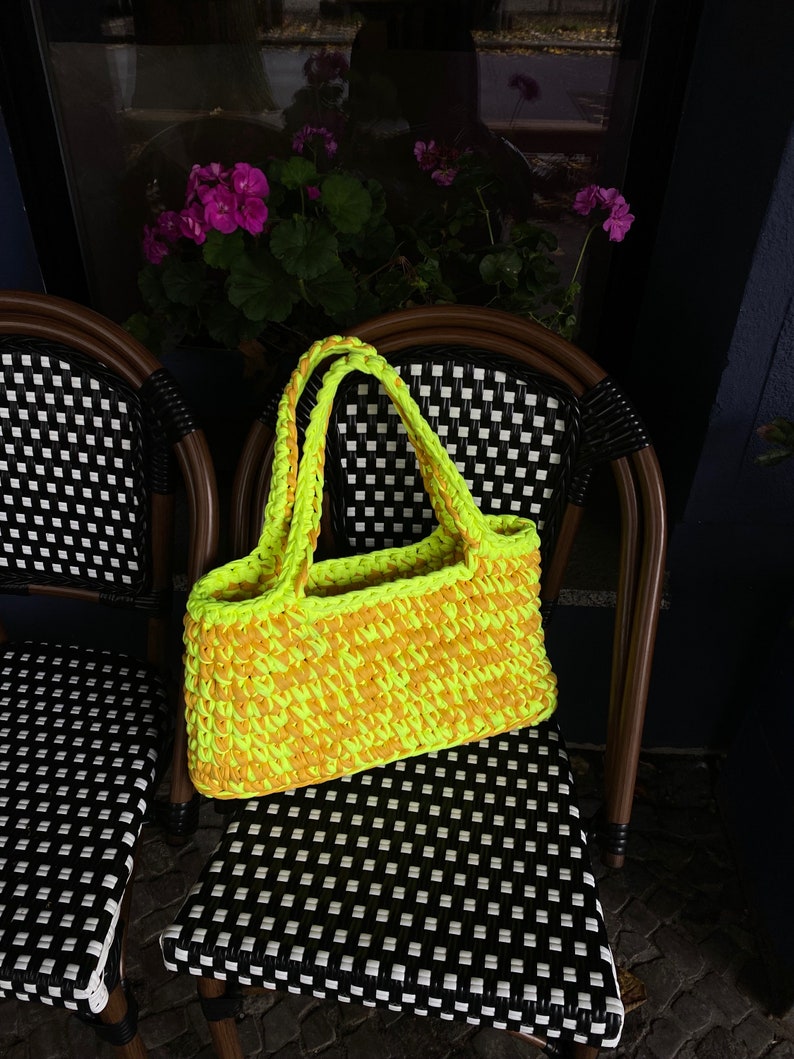 May include: A bright yellow and orange crocheted tote bag with thick straps. The bag is sitting on a black and white checkered chair.