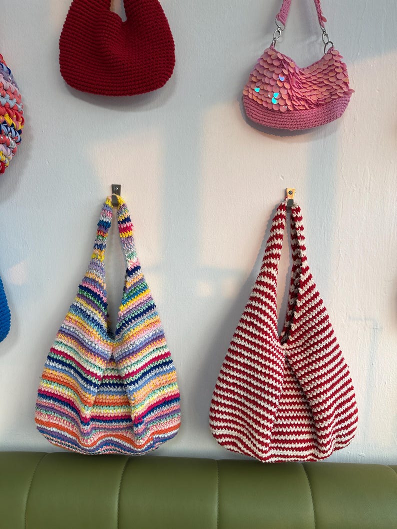 K&ouml;nnte beinhalten: Eine Sammlung handgefertigter H&auml;keltaschen in verschiedenen Farben und Stilen. Eine Tasche hat horizontale Streifen in Rot und Wei&szlig;. Eine andere Tasche hat einen Regenbogen aus Farben. Weitere Taschen sind eine rote Tasche und eine pinkfarbene Paillettentasche.