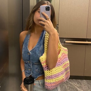 May include: A woman wearing a denim vest and blue jeans is holding a yellow, pink, and purple striped crochet tote bag. She is also holding a stainless steel travel mug.