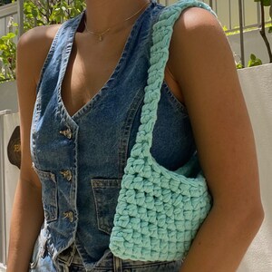 May include: A light blue crocheted bag with a single strap. The bag is worn over the shoulder of a person wearing a denim vest.