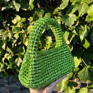 Pu&ograve; includere: Una borsa a mano verde brillante, tessuta a mano, con una forma rettangolare strutturata e un manico arrotondato. La borsa &egrave; realizzata in un materiale lucido e strutturato, su uno sfondo di fogliame verde e un muro di mattoni.