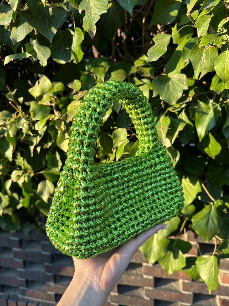 Pu&ograve; includere: Una borsa a mano verde brillante, tessuta a mano, con una forma rettangolare strutturata e un manico arrotondato. La borsa &egrave; realizzata in un materiale lucido e strutturato, su uno sfondo di fogliame verde e un muro di mattoni.