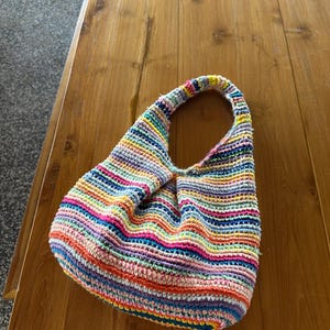 K&ouml;nnte beinhalten: Eine handgefertigte H&auml;keltasche mit einem einzelnen Tr&auml;ger. Die Tasche hat horizontale Streifen in verschiedenen Farben, darunter Rosa, Blau, Gelb und Orange. Die Tasche liegt auf einem Holztisch.