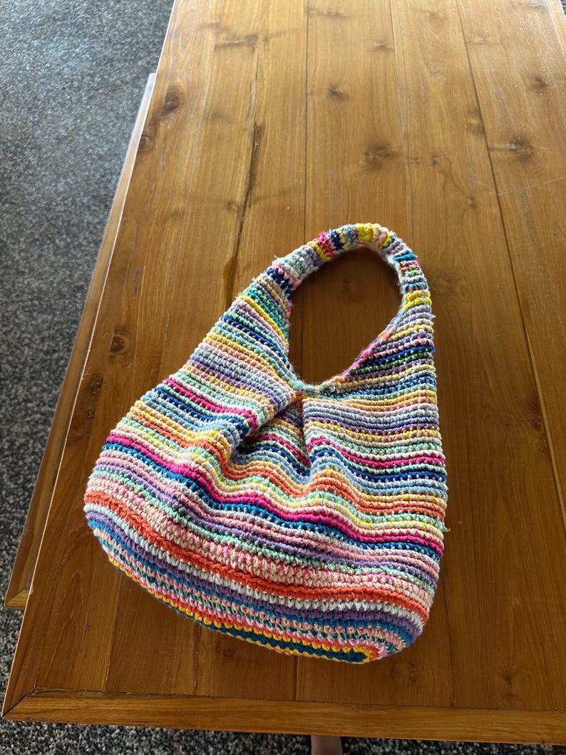K&ouml;nnte beinhalten: Eine handgefertigte H&auml;keltasche mit einem einzelnen Tr&auml;ger. Die Tasche hat horizontale Streifen in verschiedenen Farben, darunter Rosa, Blau, Gelb und Orange. Die Tasche liegt auf einem Holztisch.