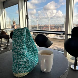Pu&ograve; includere: Una borsa di uncinetto verde acqua e bianca si trova su un tavolo grigio accanto a una tazza di caff&egrave; bianca. La tazza &egrave; piena di caff&egrave; e si vede una vista panoramica della citt&agrave; attraverso una finestra sullo sfondo.