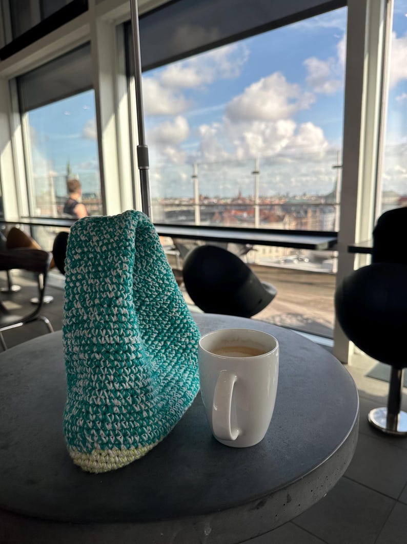 Pu&ograve; includere: Una borsa di uncinetto verde acqua e bianca si trova su un tavolo grigio accanto a una tazza di caff&egrave; bianca. La tazza &egrave; piena di caff&egrave; e si vede una vista panoramica della citt&agrave; attraverso una finestra sullo sfondo.